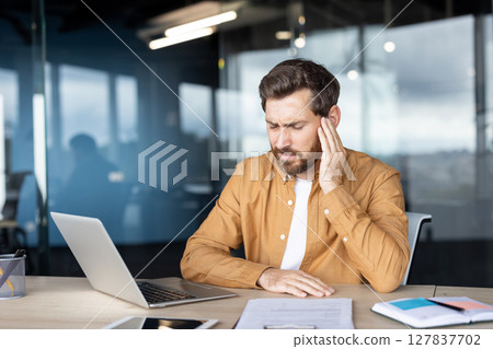 A man, experiencing ear pain, sits at a desk in an office. He holds his head and looks unwell while working on a laptop 127837702