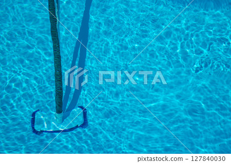 Manual pool vacuum with flexible hose and telescopic pole cleaning the bottom of a clear swimming pool during regular maintenance in bright sunlight. 127840030