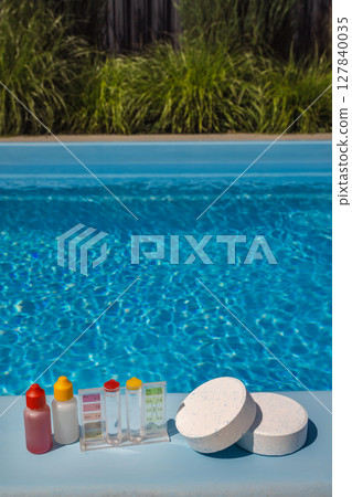 Poolside scene with chlorine tablets, water test kit, and reagent bottles used for testing and maintaining proper chemical levels in a backyard swimming pool. 127840035