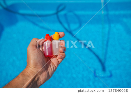 Close-up of a hand holding two dropper bottles with red and yellow caps, used for pool water testing, above a clean blue swimming pool in sunlight. 127840038