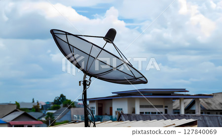 black satellite dish mounted on the roof of a building. The antenna is pointed upward, against a clear blue sky with fluffy white clouds. The design of the antenna includes a mesh dish and an elongate black satellite dish mounted on the roof of a building. The antenna is pointed upward, against a clear blue sky with fluffy white clouds. The design of the antenna includes a mesh dish and an elongate 127840069