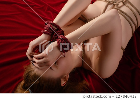 Topless woman in lingerie bound with shibari ropes on red velvet surface Topless woman in lingerie bound with shibari ropes on red velvet surface 127840412