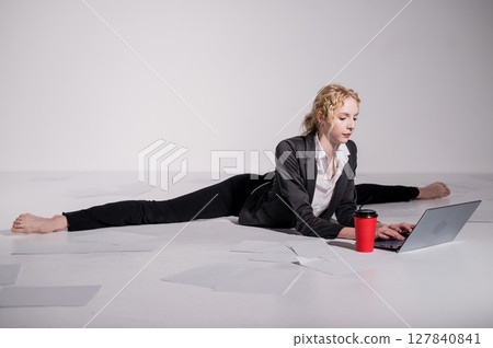 A ballerina dressed in a business suit sits in a middle twine, works at a laptop and drinks coffee. Flexible woman works at the computer A ballerina dressed in a business suit sits in a middle twine, works at a laptop and drinks coffee. Flexible woman works at the computer 127840841