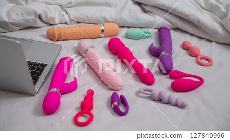 Collection of sex toys on a white sheet. Various vibrators and a laptop on the bed.  127840996