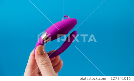 A woman holds a curved sex toy in lubrication on a blue background. Vaginally clitoral vibrator.  127841015