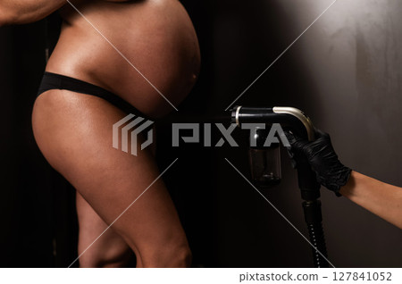 Close-up of the belly of a pregnant woman undergoing an instant tanning procedure. The master applies bronzer with a spray. Close-up of the belly of a pregnant woman undergoing an instant tanning procedure. The master applies bronzer with a spray. 127841052