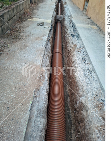 Workers are preparing a long ditch in an urban environment where a sewerage pipe is being laid. The ditch is lined with soil and gravel, indicating ongoing infrastructure work. 127841806