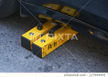 Close-Up of Car Wheel Secured with Yellow Wheel Clamp on Pavement Close-Up of Car Wheel Secured with Yellow Wheel Clamp on Pavement 127844403