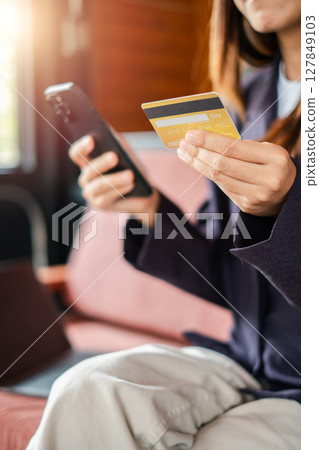 Woman using smartphone and credit card for online shopping, seated in a cozy room with sunlight streaming in. Woman using smartphone and credit card for online shopping, seated in a cozy room with sunlight streaming in. 127849103