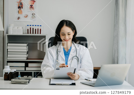Confident young  Asian female doctor in white medical uniform sit at desk working on computer. Smiling use laptop write in medical journal 127849179