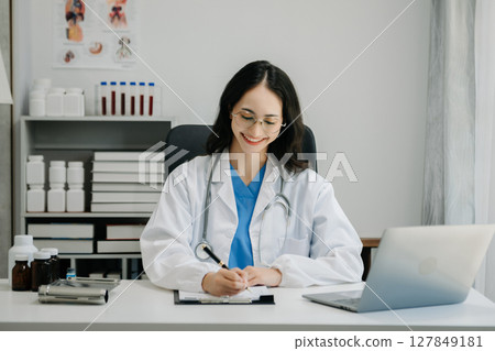 Confident young Asian female doctor in white medical uniform sit at desk working on computer. Smiling use laptop write in medical journal Confident young Asian female doctor in white medical uniform sit at desk working on computer. Smiling use laptop write in medical journal 127849181