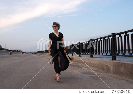 Woman in wrap dress walks by railing on urban seafront. Woman in wrap dress walks by railing on urban seafront. 127850648