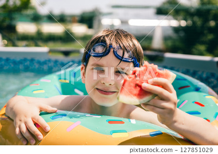 Smiling boy in pool float showing watermelon slice on a sunny summer day at the backyard Smiling boy in pool float showing watermelon slice on a sunny summer day at the backyard 127851029