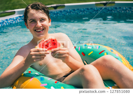 Teen boy eating juicy watermelon while relaxing in colorful pool float on a sunny summer day Teen boy eating juicy watermelon while relaxing in colorful pool float on a sunny summer day 127851080