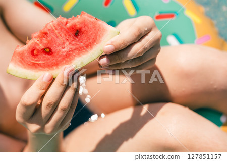 Child hands holding juicy watermelon slice on inflatable float during summer pool day Child hands holding juicy watermelon slice on inflatable float during summer pool day 127851157