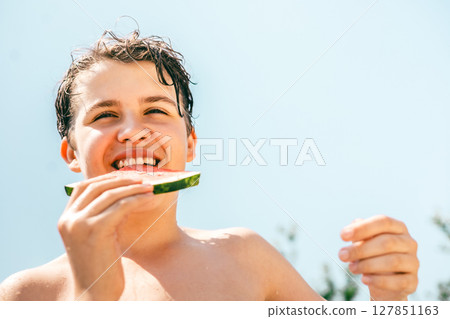 Smiling teen boy enjoying fresh watermelon outdoors on a sunny summer day under blue sky Smiling teen boy enjoying fresh watermelon outdoors on a sunny summer day under blue sky 127851163