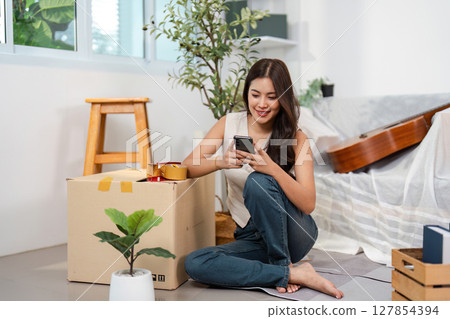 Embracing Change. A woman sits on the floor with her phone, surrounded by moving boxes and plants. 127854394