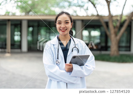 Doctor smiling confidently outdoors with tablet in hand. Healthcare professional in modern setting. Doctor smiling confidently outdoors with tablet in hand. Healthcare professional in modern setting. 127854520