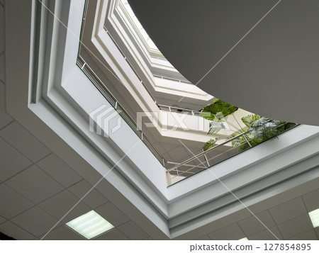 Modern multi level office atrium with geometric architectural lines, white ceilings, glass railings, and tropical plants creating bright and spacious business interior Modern multi level office atrium with geometric architectural lines, white ceilings, glass railings, and tropical plants creating bright and spacious business interior 127854895