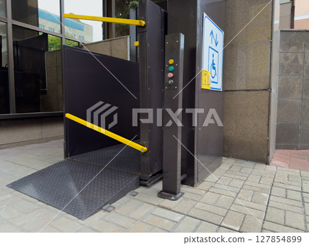 Modern vertical lift for people with disabilities near office building entrance, equipped with push buttons, safety bars, and accessibility signage to ensure inclusive access for wheelchair users Modern vertical lift for people with disabilities near office building entrance, equipped with push buttons, safety bars, and accessibility signage to ensure inclusive access for wheelchair users 127854899