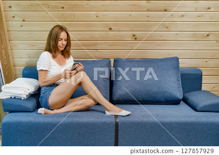 Woman in a T-shirt and shorts sitting on the sofa with a smartphone in her hands Woman in a T-shirt and shorts sitting on the sofa with a smartphone in her hands 127857929