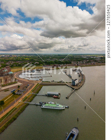 An aerial view of a modern marina with yachts docked along curved piers. Surrounding the harbor are residential buildings with green rooftops, open squares, and parking areas near the waterfront. 127858425