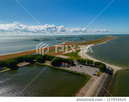 Aerial view of a narrow dam or causeway flanked by expansive water bodies on both sides, leading toward the horizon under a vibrant blue sky with scattered clouds. 127858426