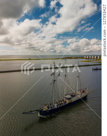A majestic sailing ship glides across calm waters under a partly cloudy sky, with coastal landscapes and a modern dam structure visible in the background. 127858427