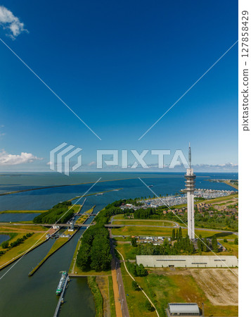 An aerial view of a coastal town with a harbor, colorful buildings, a tall communication tower, and a replica ship docked along the water. Surrounding areas include parking lots and green spaces. 127858429