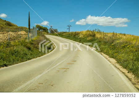 The asphalt road goes into a curve running among fields and meadows on a sunny summer day. Blue sky with white curly clouds. Good weather. Travel. 127859352