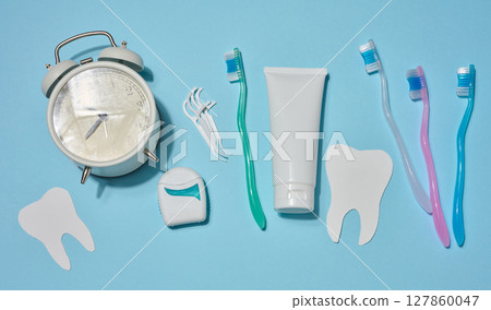 Set of items for oral hygiene, white tube of toothpaste, toothbrushes, dental floss and white alarm clock, and decorative paper teeth, on blue background. Top view. Set of items for oral hygiene, white tube of toothpaste, toothbrushes, dental floss and white alarm clock, and decorative paper teeth, on blue background. Top view. 127860047