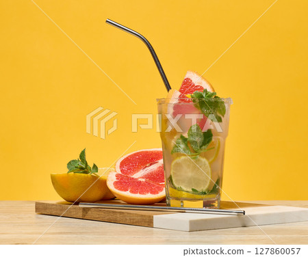 Glass with cocktail and grapefruit, lemon and orange slices, metal straw inside. Yellow background 127860057