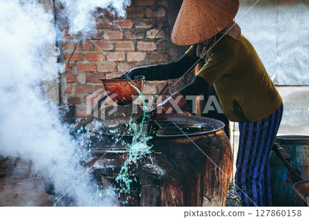 Vietnamese female worker puts out fire in furnace in reed dyeing workshop at a factory in a village. Asian woman does dirty hard men's work in a factory in Asia 127860158