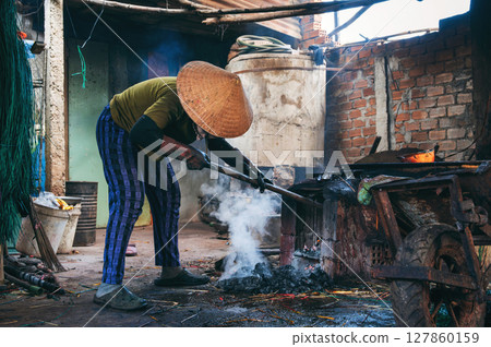 Vietnamese female stoker worker works in a workshop and lights a coal furnace at reed dyeing plant in Vietnam. Woman does dirty hard men's work in factory in Asia Vietnamese female stoker worker works in a workshop and lights a coal furnace at reed dyeing plant in Vietnam. Woman does dirty hard men's work in factory in Asia 127860159