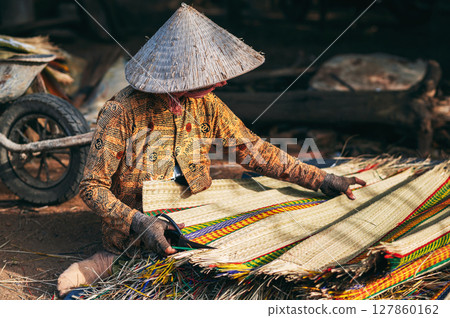 Vietnamese old woman worker in traditional straw hat Non La works at a reed mat factory in craft village in Vietnam Vietnamese old woman worker in traditional straw hat Non La works at a reed mat factory in craft village in Vietnam 127860162