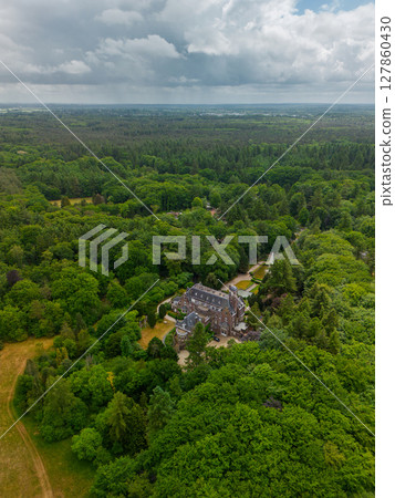 An aerial view of a historic mansion surrounded by lush green trees and a beautifully landscaped garden with a fountain, walking paths, and parking area. The scene shows vibrant summer greenery. 127860430