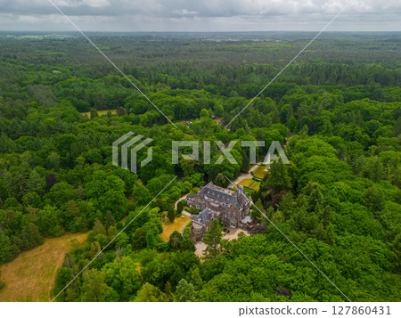 An aerial view of a historic mansion surrounded by lush green trees and a beautifully landscaped garden with a fountain, walking paths, and parking area. The scene shows vibrant summer greenery. An aerial view of a historic mansion surrounded by lush green trees and a beautifully landscaped garden with a fountain, walking paths, and parking area. The scene shows vibrant summer greenery. 127860431