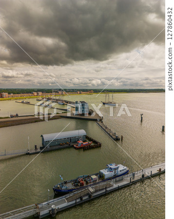 Aerial view of a harbor with several boats and ships on a large body of water under dramatic, cloudy skies, with a modern waterfront building and docks visible in the foreground. Aerial view of a harbor with several boats and ships on a large body of water under dramatic, cloudy skies, with a modern waterfront building and docks visible in the foreground. 127860432