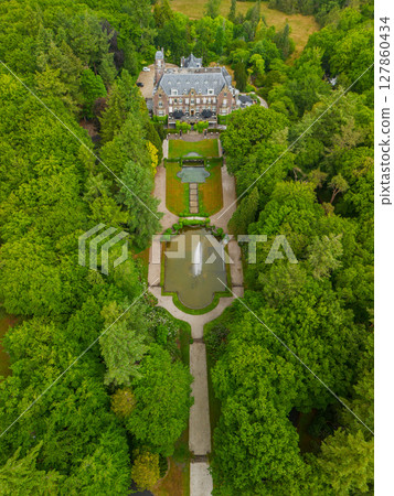 An aerial view of a historic mansion surrounded by lush green trees and a beautifully landscaped garden with a fountain, walking paths, and parking area. The scene shows vibrant summer greenery. An aerial view of a historic mansion surrounded by lush green trees and a beautifully landscaped garden with a fountain, walking paths, and parking area. The scene shows vibrant summer greenery. 127860434