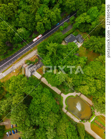 A drone view of a road cutting through dense green forest, with open grassy fields on one side and residential houses tucked among the trees, capturing a peaceful blend of nature and infrastructure. 127860436