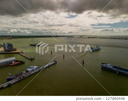 Aerial view of a harbor with several boats and ships on a large body of water under dramatic, cloudy skies, with a modern waterfront building and docks visible in the foreground. Aerial view of a harbor with several boats and ships on a large body of water under dramatic, cloudy skies, with a modern waterfront building and docks visible in the foreground. 127860446