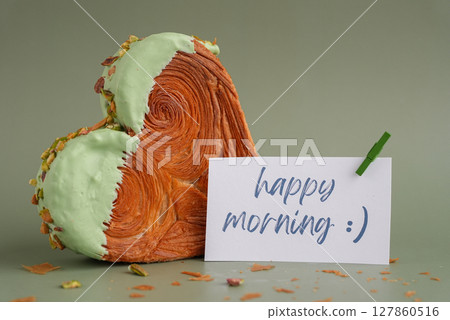 HAPPY MORNING caption on paper note love symbol Heart shaped pistachio croissant on green background with copy space. Puff pastry trendy sweet dessert bakery. Creamy dough experiment HAPPY MORNING caption on paper note love symbol Heart shaped pistachio croissant on green background with copy space. Puff pastry trendy sweet dessert bakery. Creamy dough experiment 127860516
