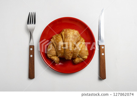 Spoon and fork with freshly baked croissant on a red plate with a white background. Top view Spoon and fork with freshly baked croissant on a red plate with a white background. Top view 127862074