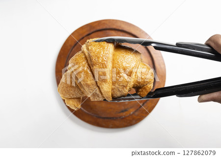 Tongs with freshly baked croissants on a wooden tray with a white background. Top view Tongs with freshly baked croissants on a wooden tray with a white background. Top view 127862079