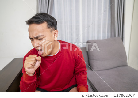 Indonesian southeast asian man sitting on the couch at home and coughing. Suffers from an attack of asthma or allergies. Healthcare concept 127862307
