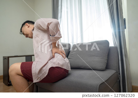 Indonesian southeast asian man suffering from back pain while sitting on the living room couch at home. Healthcare concept 127862320