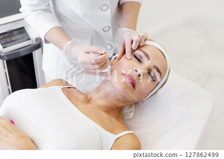 Mature woman getting rejuvenation injection in beauty clinic by cosmetologist in protective gloves and white coat, anti-aging facial treatment to smooth wrinkles and restore youthful skin appearance Mature woman getting rejuvenation injection in beauty clinic by cosmetologist in protective gloves and white coat, anti-aging facial treatment to smooth wrinkles and restore youthful skin appearance 127862499