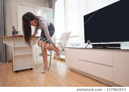 Indonesian southeast asian businesswoman feels an ache because of wearing high heels for too long in a hybrid workplace. Laptop and paperwork on the table. Business concept. Remote working from home 127862653