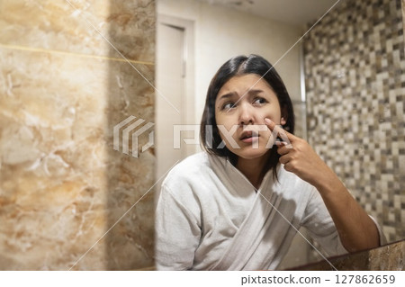 Indonesian southeast asian woman wrapped in a towel, looking at herself in the mirror while squeezing an acne spot. Natural skincare and wellness concept 127862659