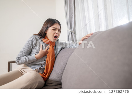 Indonesian southeast asian woman in a scarf sitting on the couch at home and coughing. Suffer from fever, flu, and cold. Healthcare concept Indonesian southeast asian woman in a scarf sitting on the couch at home and coughing. Suffer from fever, flu, and cold. Healthcare concept 127862776